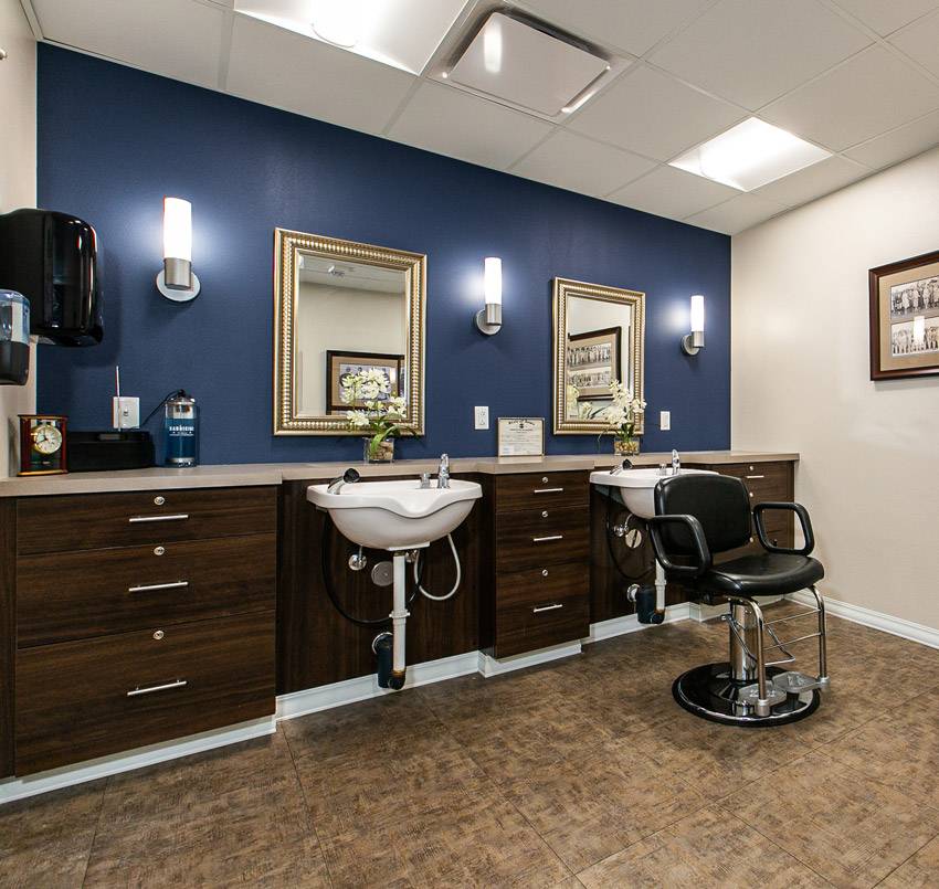 Hawkeye Care Center salon area with a dark blue wall, dark wood cabinets, two salon sinks, a salon chair, and two gold-colored framed mirrors.