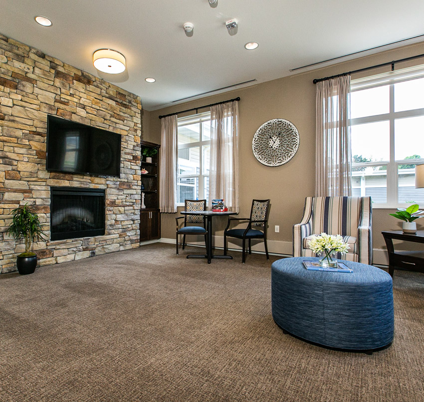 Hawkeye Care Center Living area with a TV placed over a fireplace on a brick wall, small table with two chairs, a striped arm chair, and blue round foot rest